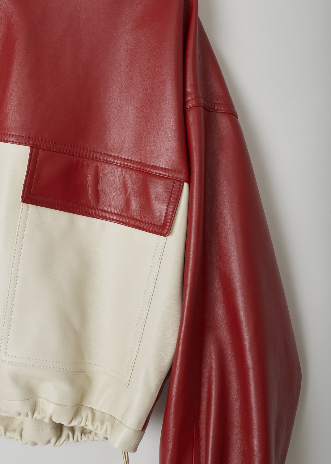 CELINE, TWO-TONE CROPPED LEATHER JACKET, 150T_2EE13_27LV, Red, Beige, Detail 1, This cropped leather jacket is  two-toned, with the top half in lipstick red and the bottom half and the back in off-white. The jacket has a stand-up collar, two patch pockets with flap and a zipper in a matching off-white color. The gold-tone pull-tab is shaped in the brand's logo. The long sleeves have elasticated cuffs. The elasticated hemline has gold-tone cord locks on the inside. The jackets has a single inner pocket

