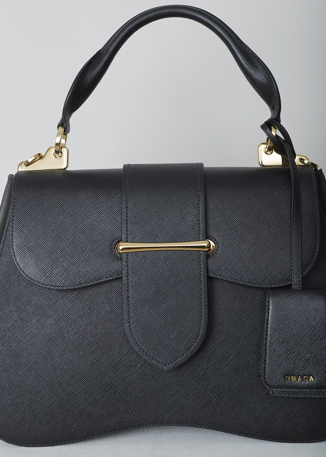 PRADA, LARGE SIDONIE TOP HANDLE BAG IN BLACK, CARTELLA_1BN002_F0002_NERO, Black, Detail, This large black Sidonie Top Handle Bag is made with Saffiano leather. The bag comes with a leather handle and an adjustable and detachable shoulder strap. The bag has gold-tone metal hardware with the brand's logo on the back. The removable leather keychain has that same gold-tone logo on it. The slip-through closure opens up to reveal its red interior. The bag has a single, spacious compartment with a zipped inner pocket to one side and a patch pocket on the other side.  

