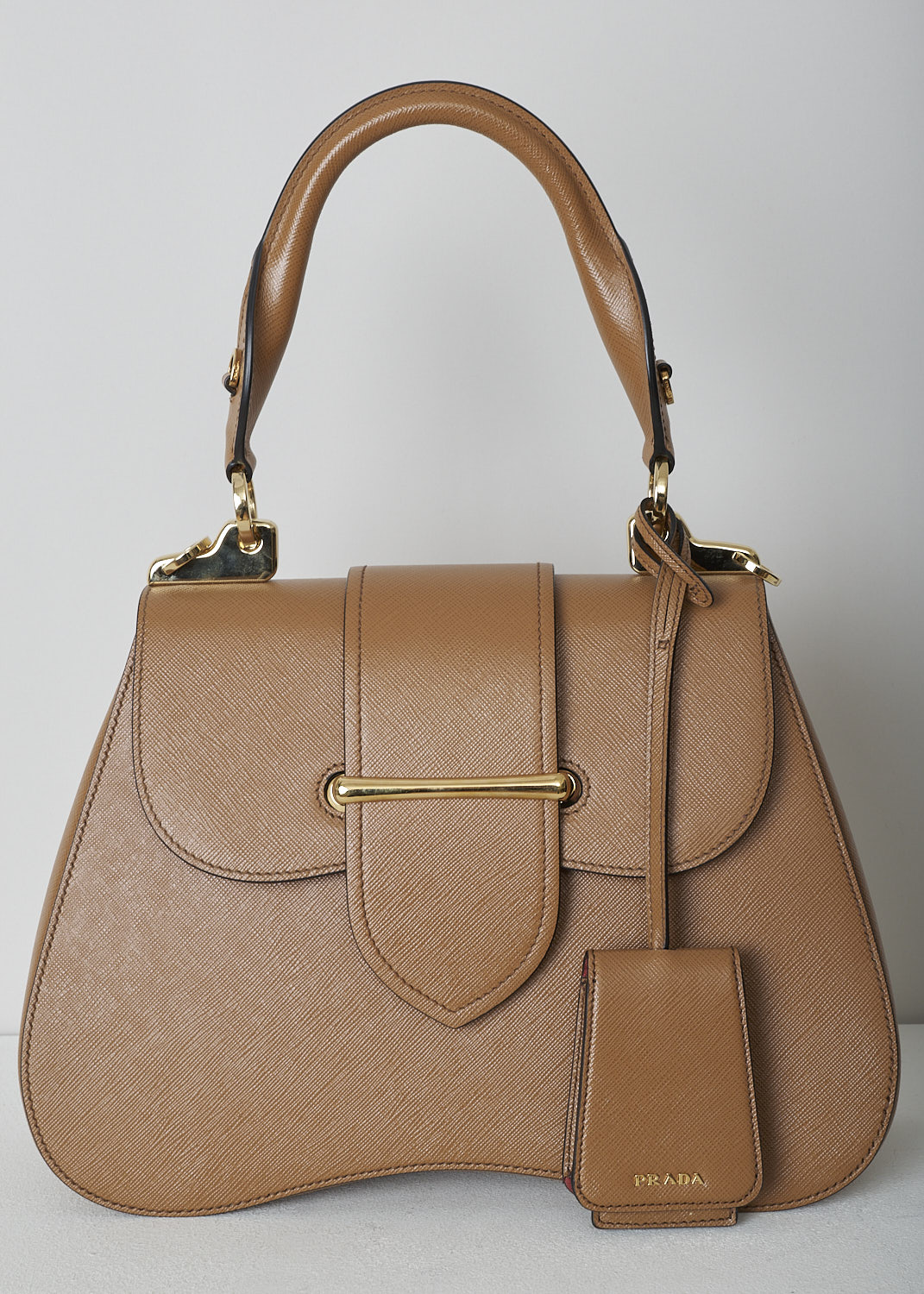 PRADA, MEDIUM SIDONIE TOP HANDLE BAG IN CARAMEL BROWN, CARTELLA_1BN005_F098L_CARAMEL, Brown, Detail, This mid-sized caramel brown Sidonie Top Handle Bag is made with Saffiano leather. The bag comes with a leather handle and an adjustable and detachable shoulder strap. The bag has gold-tone metal hardware with the brand's logo on the back. The removable leather keychain has that same gold-tone logo on it. The slip-through closure opens up to reveal its red interior. The bag has a single, spacious compartment with a zipped inner pocket to one side and a patch pocket on the other side.  

