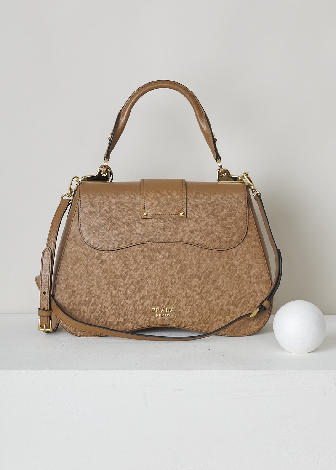 PRADA, CARAMEL BROWN SIDONIE TOP HANDLE BAG, CARTELLA_1BN002_F098L_CARAMEL, Brown, Back, This large caramel brown Sidonie Top Handle Bag is made with Saffiano leather. The bag comes with a leather handle and an adjustable and detachable shoulder strap. The bag has gold-tone metal hardware with the brand's logo on the back. The removable leather keychain has that same gold-tone logo on it. The slip-through closure opens up to reveal its red interior. The bag has a single, spacious compartment with a zipped inner pocket to one side and a patch pocket on the other side.   

