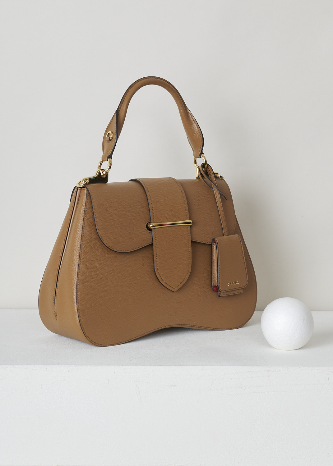 PRADA, CARAMEL BROWN SIDONIE TOP HANDLE BAG, CARTELLA_1BN002_F098L_CARAMEL, Brown, Side, This large caramel brown Sidonie Top Handle Bag is made with Saffiano leather. The bag comes with a leather handle and an adjustable and detachable shoulder strap. The bag has gold-tone metal hardware with the brand's logo on the back. The removable leather keychain has that same gold-tone logo on it. The slip-through closure opens up to reveal its red interior. The bag has a single, spacious compartment with a zipped inner pocket to one side and a patch pocket on the other side.   

