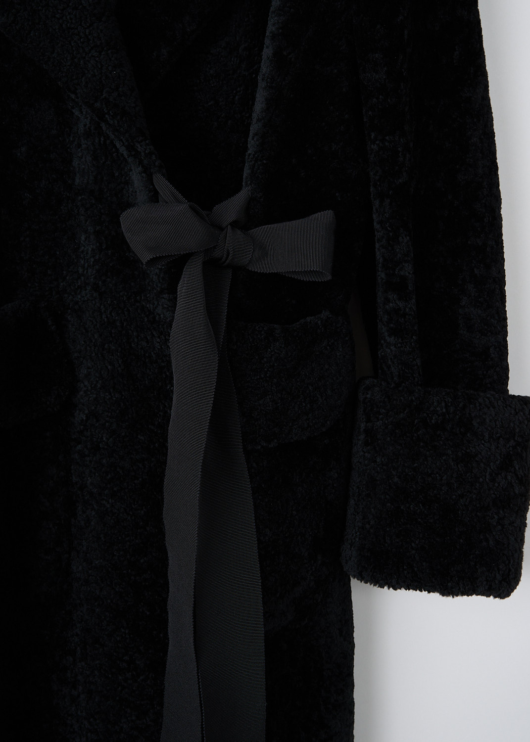 Prada, Black lammy coat with ribbon closure, 1XM2_MONTONE_56950_F0002_NERO, Black, Detail, This black lammy coat has an exaggerated notched lapel. To one side, a black grosgrain ribbon functions as the closure. The closure is supported by a concealed button on the inside. The long sleeves have broad folded cuffs. In the front, the coat has patch pockets with decorative flaps. The coat has a straight hemline.  
