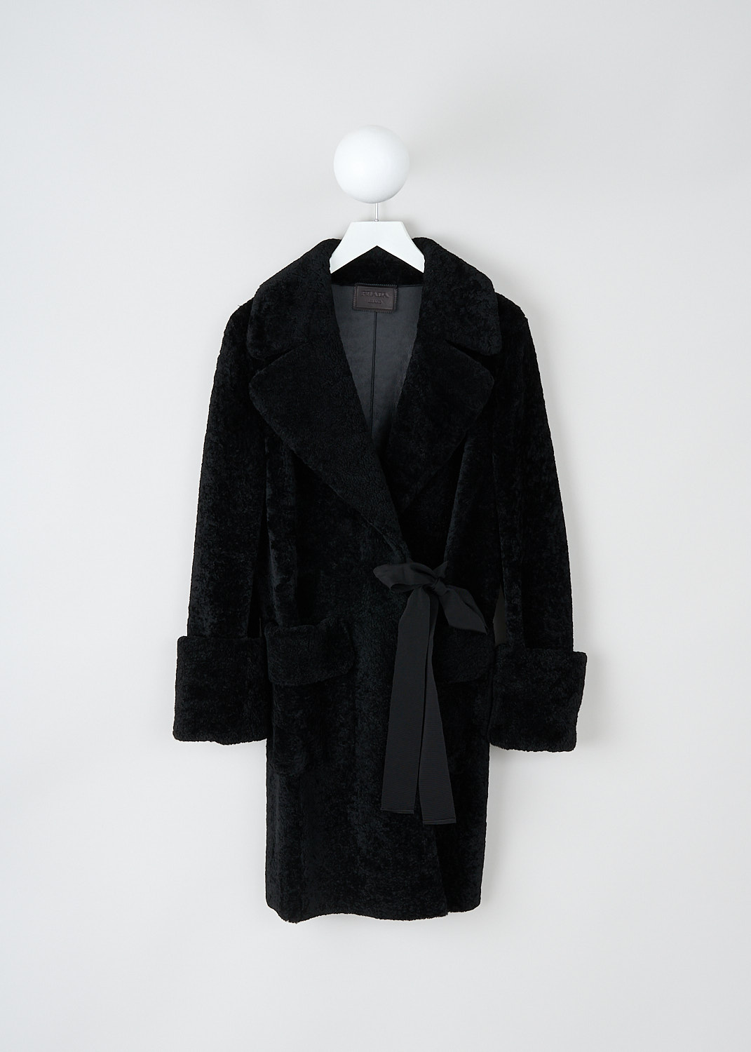 Prada, Black lammy coat with ribbon closure, 1XM2_MONTONE_56950_F0002_NERO, Black, Front, This black lammy coat has an exaggerated notched lapel. To one side, a black grosgrain ribbon functions as the closure. The closure is supported by a concealed button on the inside. The long sleeves have broad folded cuffs. In the front, the coat has patch pockets with decorative flaps. The coat has a straight hemline.  
