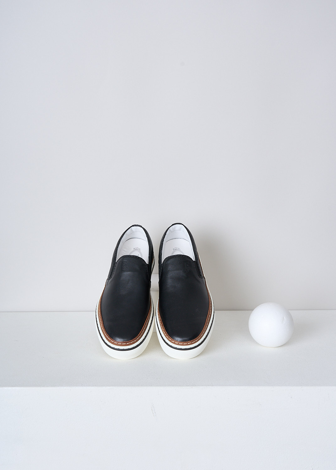 TODS, BLACK LEATHER SLIP-ON SHOES, Top, Black, Side, These slip-on shoes have a black leather top with elasticated inserts on either sides. The brand's logo can be found debossed on the front. These shoes have a broad white rubber sole with a brown, white and black trim. 
